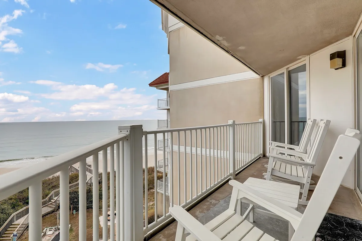 A private oceanfront balcony featuring white chairs behind a balcony railing, with expansive views of the ocean and a cloudy sky. The scene exudes calm and relaxation, perfect for quiet moments or enjoying a drink while overlooking the sea.