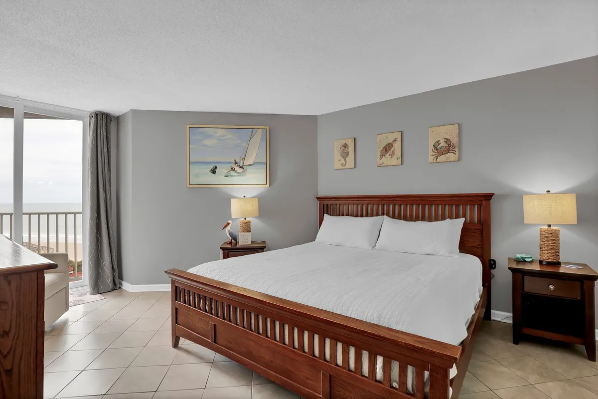 A serene master bedroom featuring a large, inviting bed positioned near a window that offers natural light and a tranquil view. The room is decorated with calming artwork, and the ambiance promises a restful atmosphere ideal for relaxation after a day at the shore.