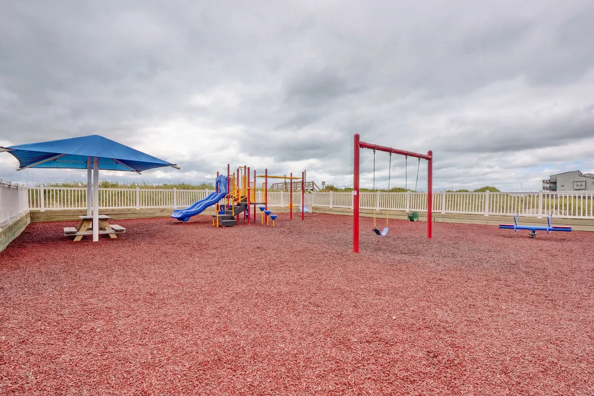 A lively playground scene, complete with various play structures, a swing set, picnic table, and shade umbrella. Children can be seen playing while adults enjoy the sun and watch from nearby, embodying energetic family fun.