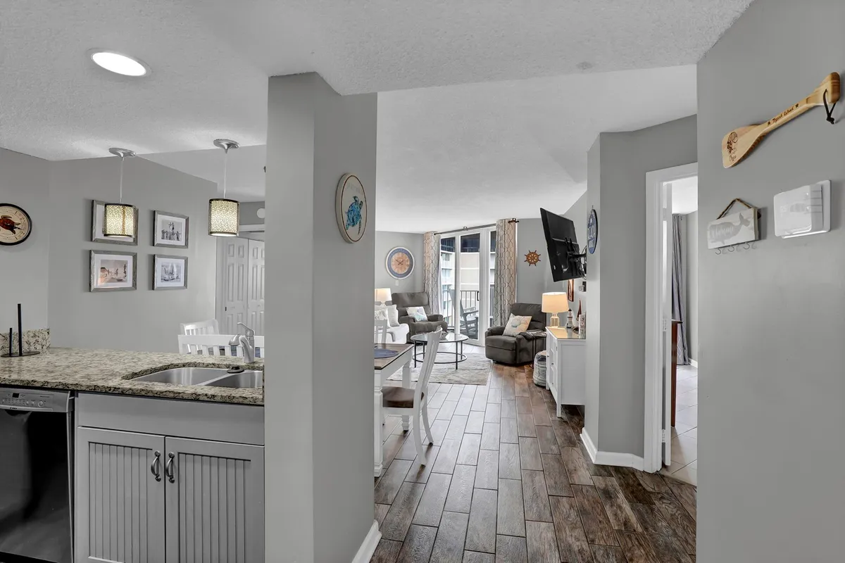 A wide-angle view from the kitchen countertop toward the living area, revealing comfortable seating, a dining table, and large windows that allow abundant sunlight and showcase the balcony. The space feels inviting and sociable, ideal for both quiet afternoons and group gatherings.