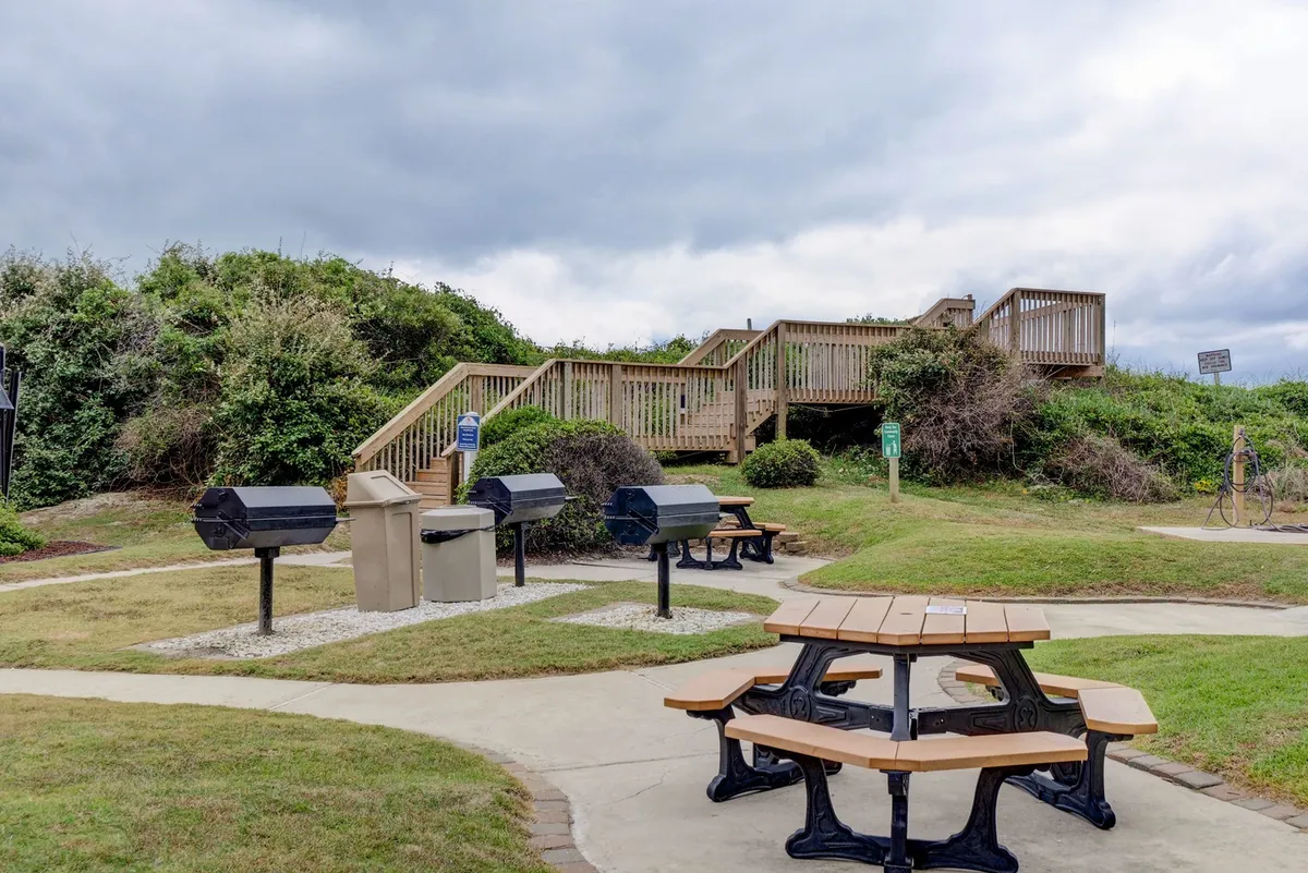 An outdoor communal area featuring picnic tables and barbecue grills, set beside a staircase leading up to a deck. The setting is open and friendly, ideal for outdoor dining and social gatherings under the sun.