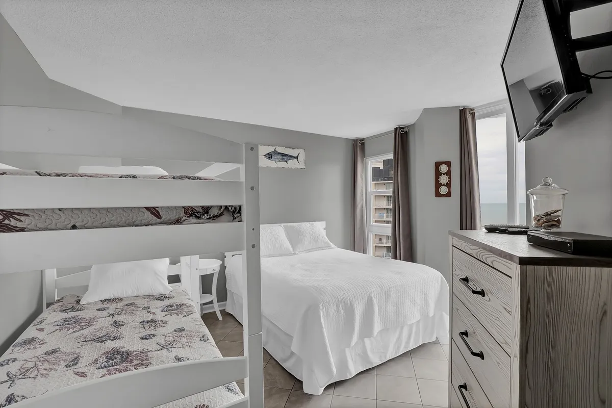 A spacious and family-friendly second bedroom with a twin-over-twin bunk bed and a cozy queen bed. The room includes a television, a window offering a lovely view, and is styled in a relaxed manner—perfect for families seeking comfort and flexibility.