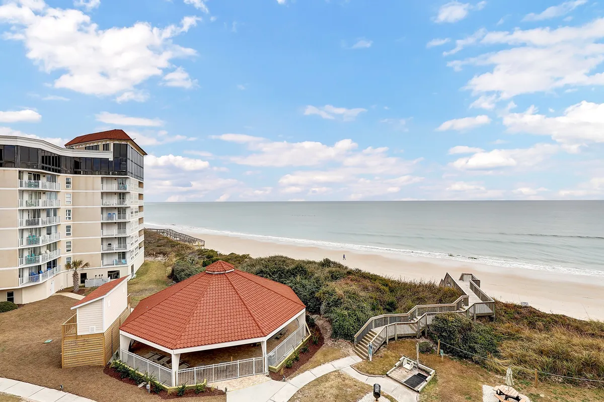 The hero image showcases the oceanfront condominium complex at North Topsail Beach, with a wide sandy beach stretching in front and a charming gazebo nestled close to the shore. The tranquil Atlantic Ocean forms a serene backdrop, while the inviting gazebo hints at spaces for relaxing or gathering with friends and family. The scene conveys a sense of peaceful coastal living, with opportunities for sunrise views, leisurely strolls on the sand, and memorable outdoor moments.
