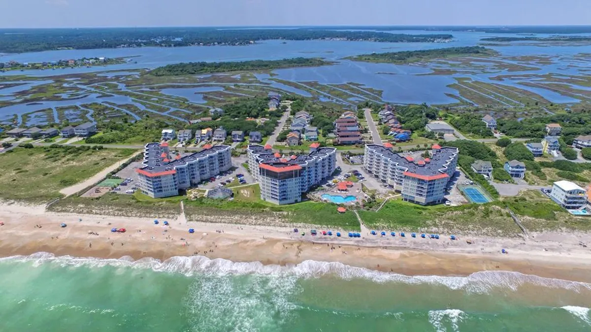 Beachfront buildings nestled close to the sand, a swimming pool in the foreground, ocean waves rolling in, and a stretch of marshland visible. The setting is ideal for swimming, sunbathing, and beach walks, with a clear connection to both the resort and the natural environment.