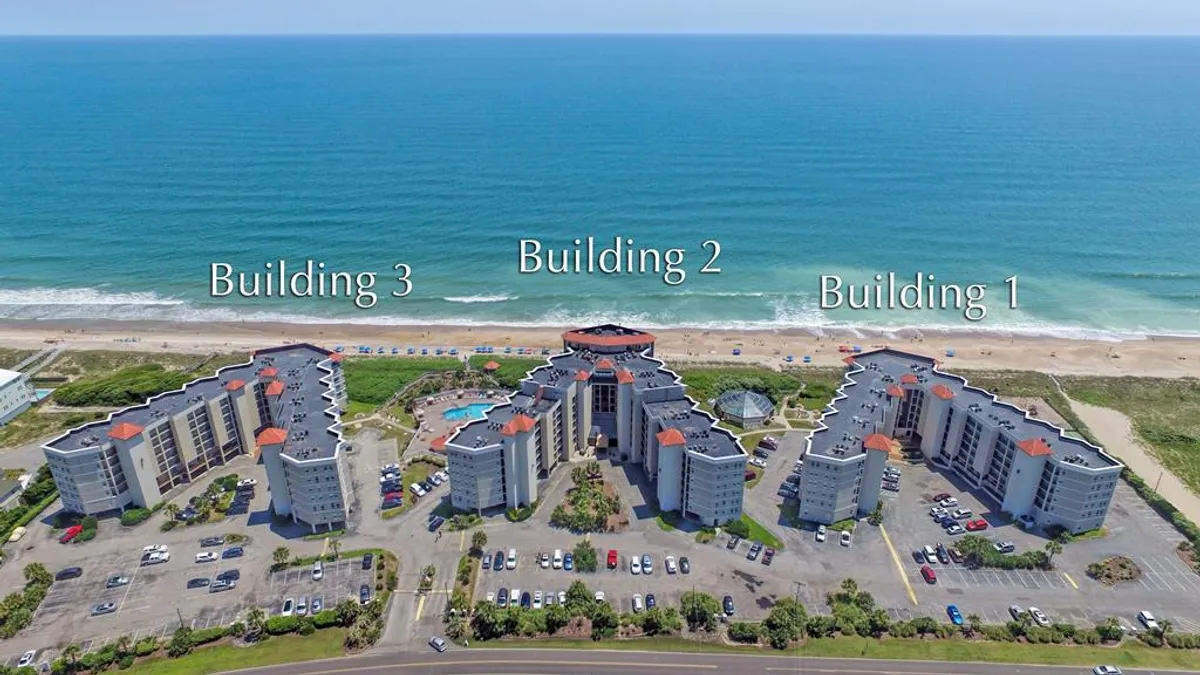 A view of the resort's buildings with a swimming pool area in the foreground and the beach visible beyond. The scene captures families relaxing, swimming, and enjoying the close proximity to both pool and ocean.