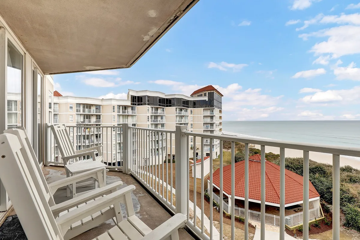 A tranquil balcony scene with white rocking chairs positioned to overlook the ocean, framed by a railing and surrounded by neighboring buildings. The space is perfect for unwinding and enjoying gentle ocean breezes, capturing the essence of private outdoor relaxation.