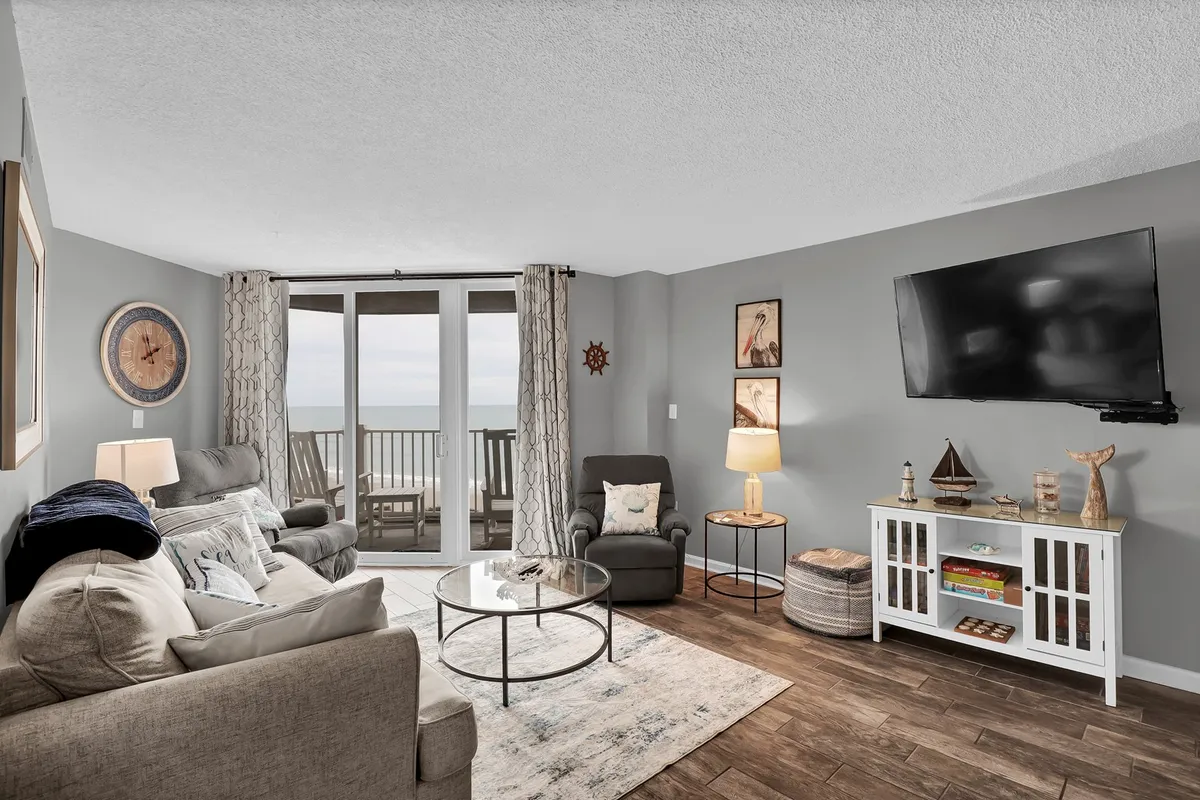 A cozy, open living room with a comfortable sofa, coffee table, large TV, and sliding glass doors that frame an inviting view of the oceanfront balcony. Natural light streams into the room, creating a tranquil, welcoming atmosphere perfect for relaxation and togetherness.