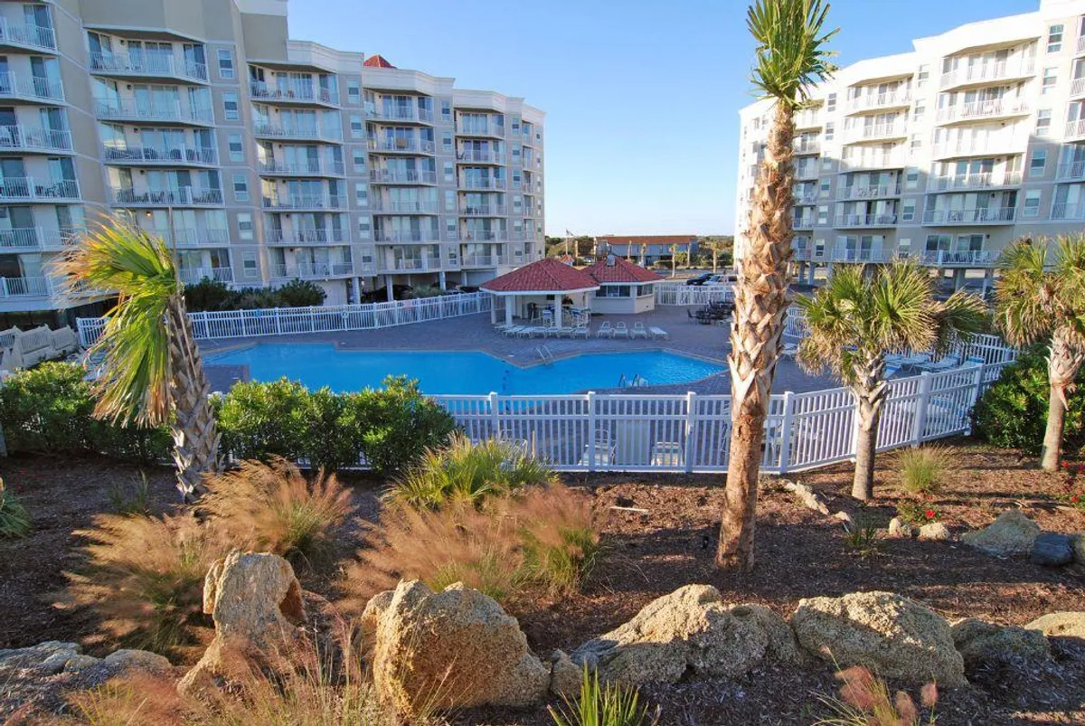 An inviting swimming pool set amidst palm trees and landscaped surroundings, with buildings visible in the background. There are areas for sunbathing and relaxing, suggesting a resort-like ambiance perfect for both peace and social gatherings.